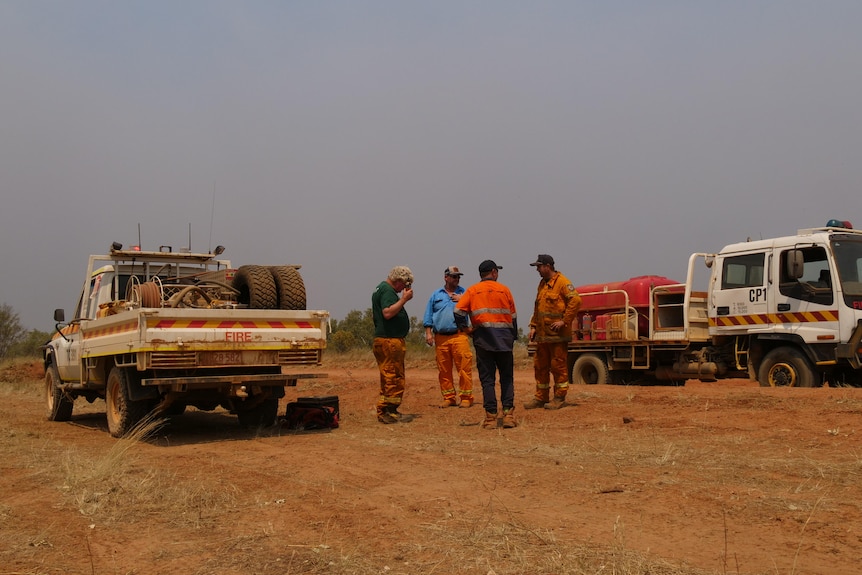 A small group of people in high vis standing on a dirt road between two parked vehicles and talking, with bushfire smoke ove