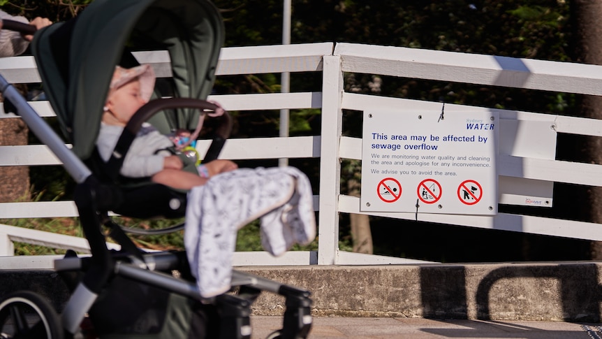A sign attached to a fence saying this area may be affected by sewage overflow next to a baby in a pram