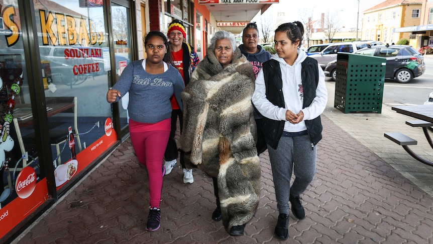 Esther Kirby OAM wrapped in her possum skin cloak walking down the street in Kerang with four women.