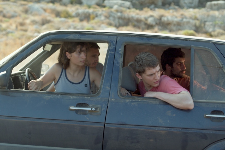 Four young people in an old battered car, they are all looking behind them, dry/desert background