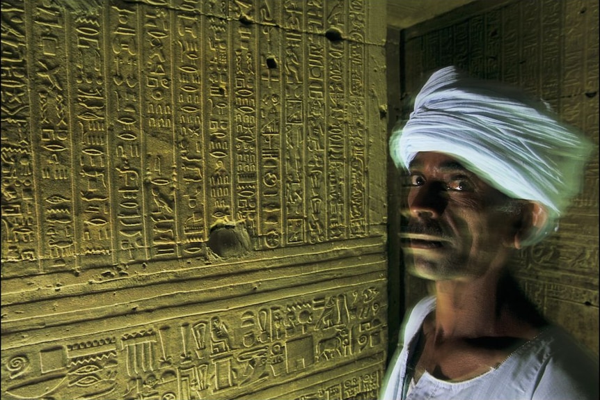 An Egyptian man stands next to hieroglyphics in a temple 