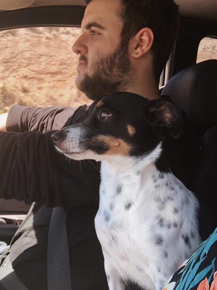 A man with dark hair, facial hair and an earring driving in a car. A small dog sits next to him.