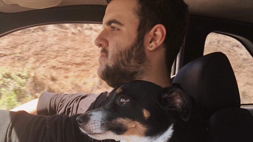 A man with dark hair, facial hair and an earring driving in a car. A small dog sits next to him.