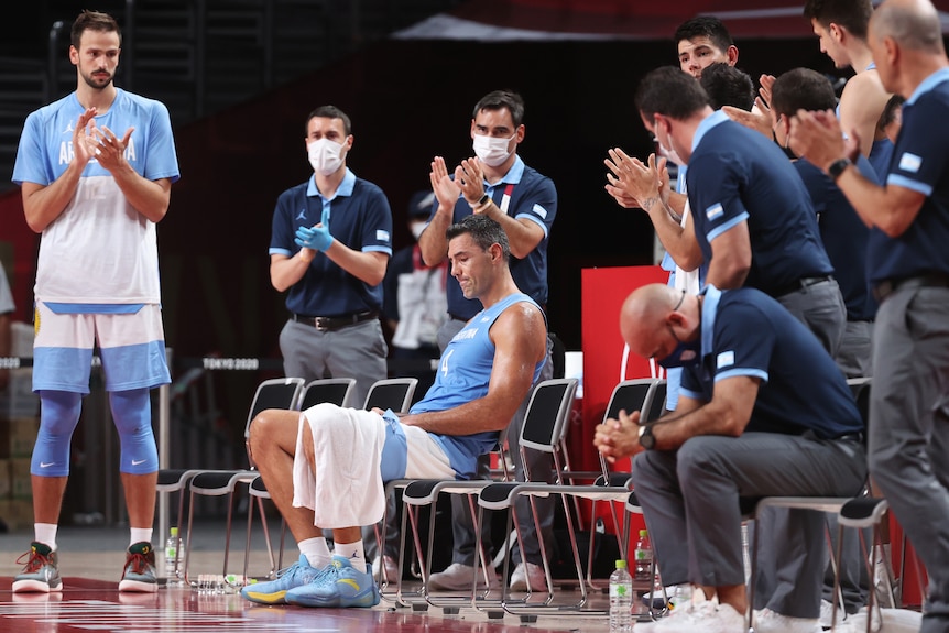 A man sits in a chair while a group of men surround him and clap