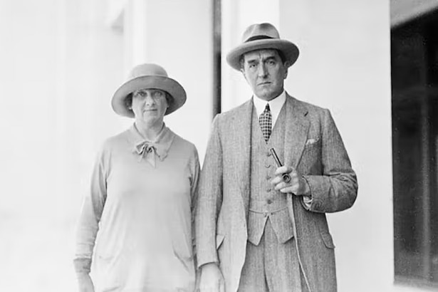 A black and white photo of a man in a three piece suit holding a cigar and his wife in a 1920s style hat