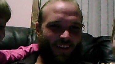 A poorly-lit shot of a young, balding man with a thick beard sitting in a living room.