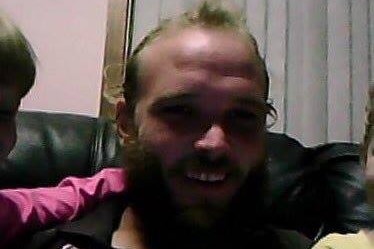 A poorly-lit shot of a young, balding man with a thick beard sitting in a living room.