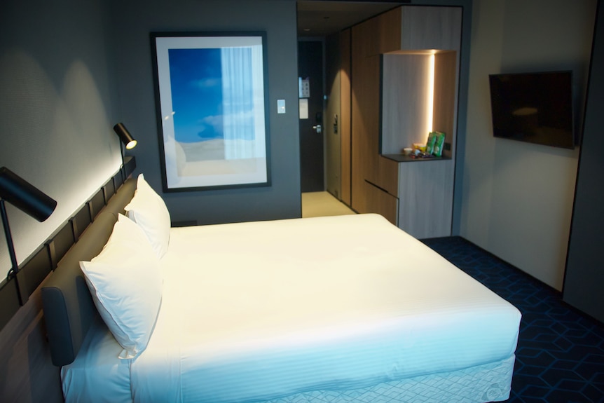 The inside of a hotel room, with a white-dressed bed and lamps on.