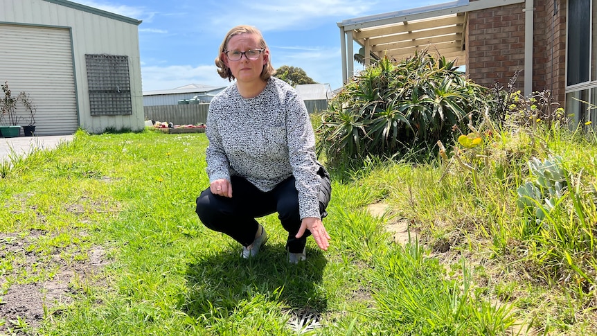 Sharon Harrison kneels on the grass in her yard to show the flood damage on her property.