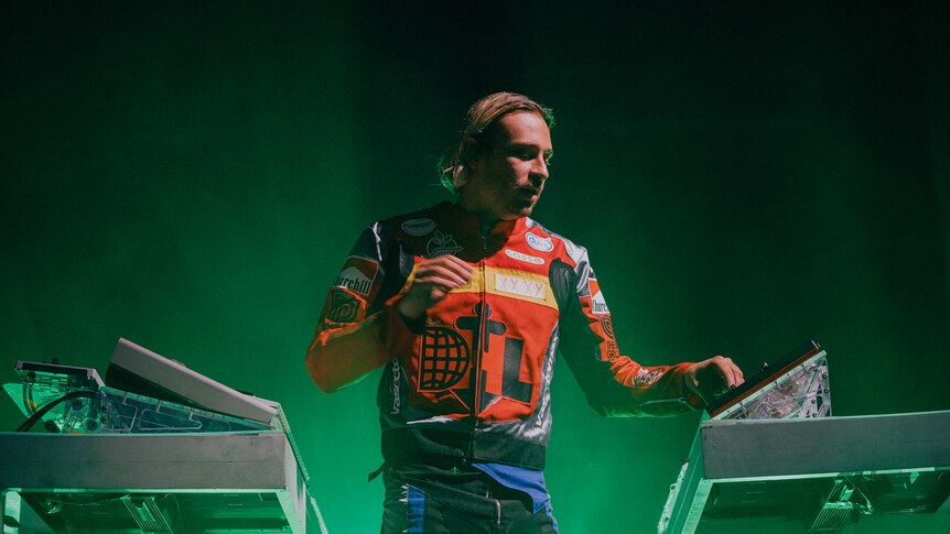 Flume wearing a motocross bike jacket and performing live between two banks of control panels illuminated by green light