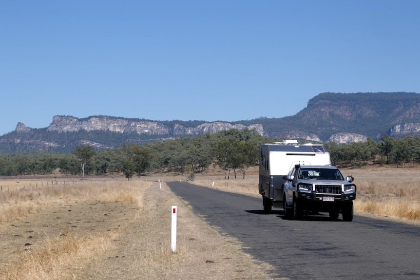 A four wheel drive towing a caravan, sandstone ranges, trees, road and grass behind.