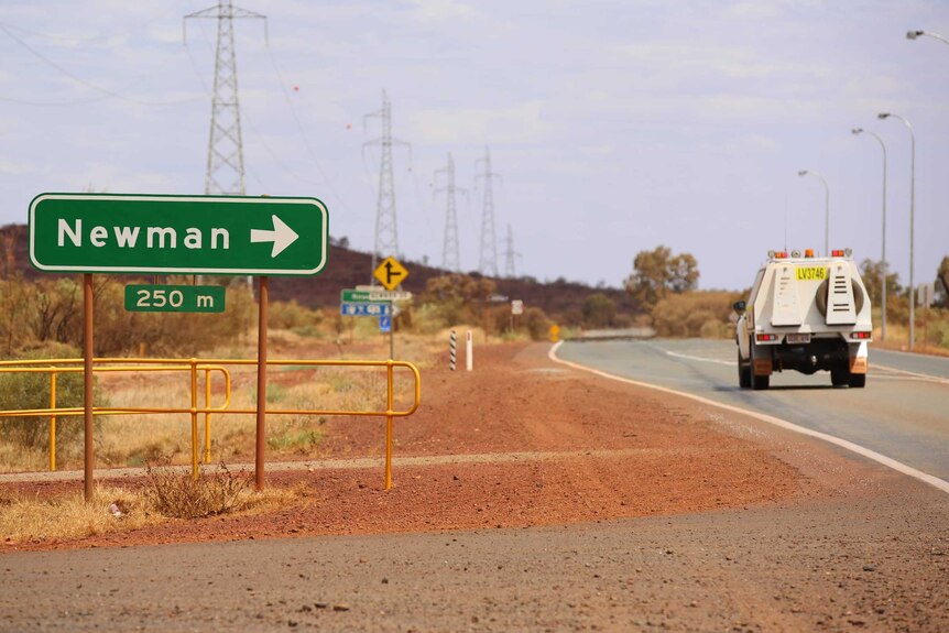 A green road sign showing Newman with an arrow point right stands next to a work vehicle on the road and a red dirt shoulder.