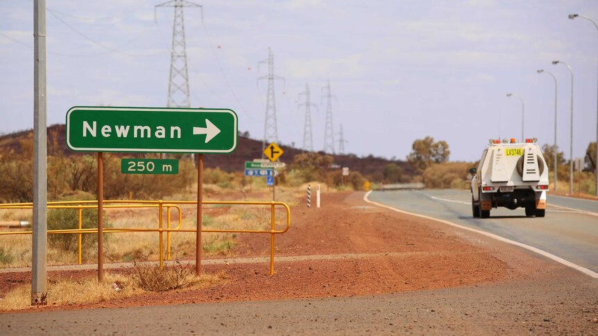 A green road sign showing Newman with an arrow point right stands next to a work vehicle on the road and a red dirt shoulder.