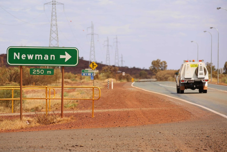 A green road sign showing Newman with an arrow point right stands next to a work vehicle on the road and a red dirt shoulder.