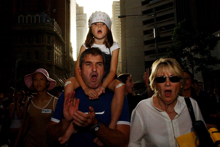Young girl sits on man's shoulders chanting as they walk through city street surrounded by protesters 