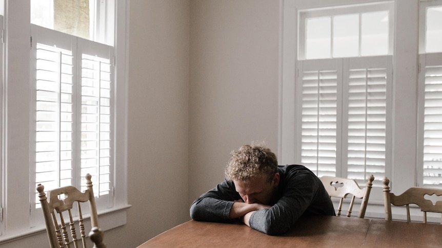 A young man slumps over with his chin resting on his arms, at a dining room table.