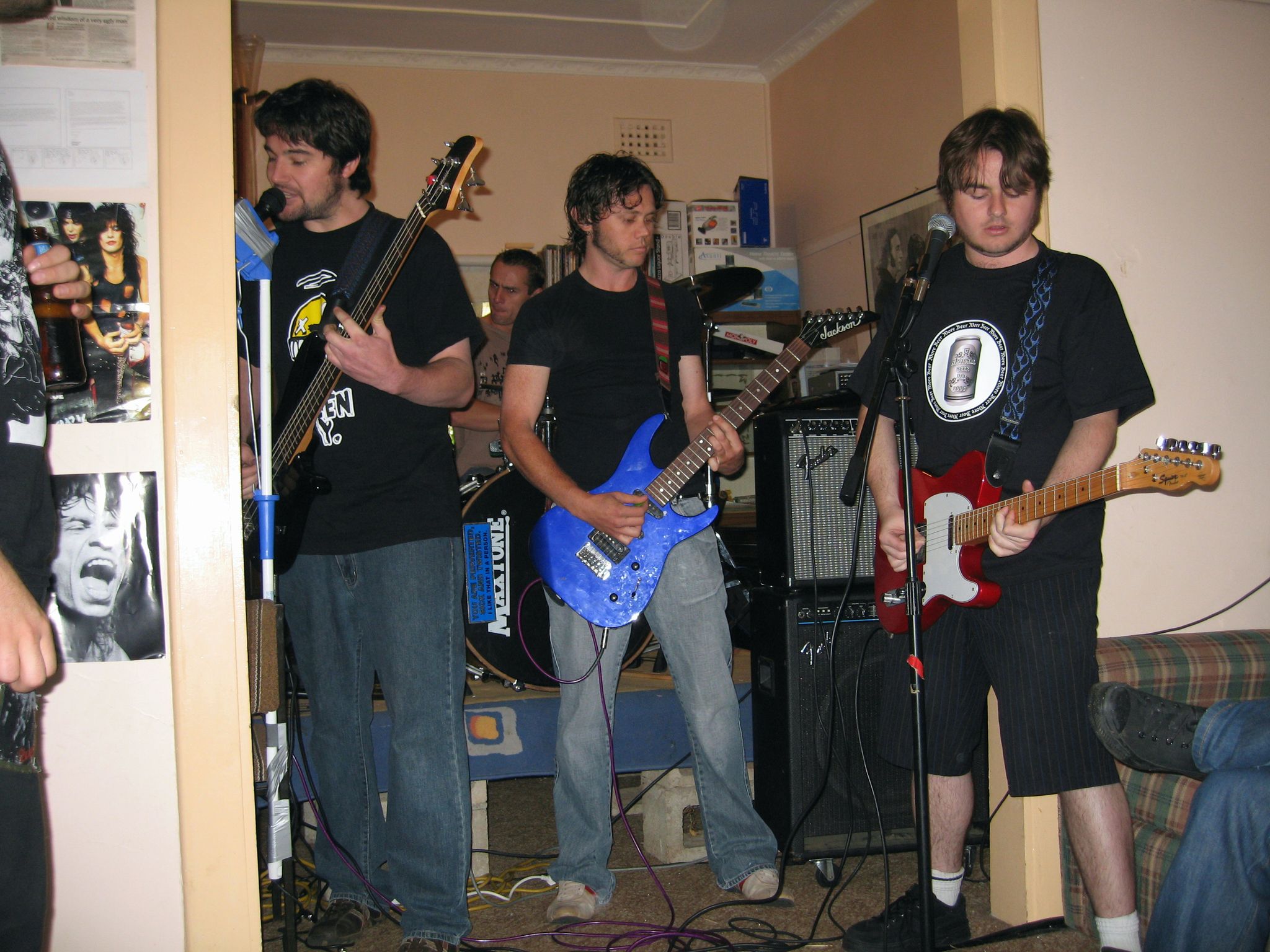 Three young men in black t-shirts with guitars and standing in front of microphones, playing in a band in a small room.