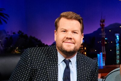 James Corden in a black suit with white sparkles smiles at the camera on the backdrop of the late late show. 