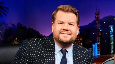 James Corden in a black suit with white sparkles smiles at the camera on the backdrop of the late late show. 