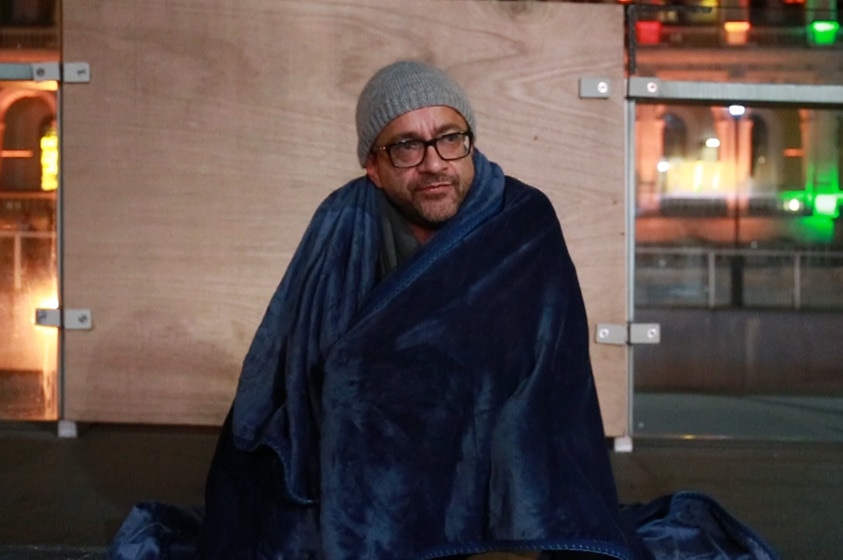 A man wrapped in a blue blanket sitting on a concrete path in Brisbane CBD.