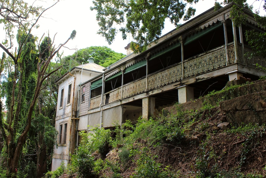 A large white and green mansion, covered in rust and decay, surrounded by old creeper-covered trees