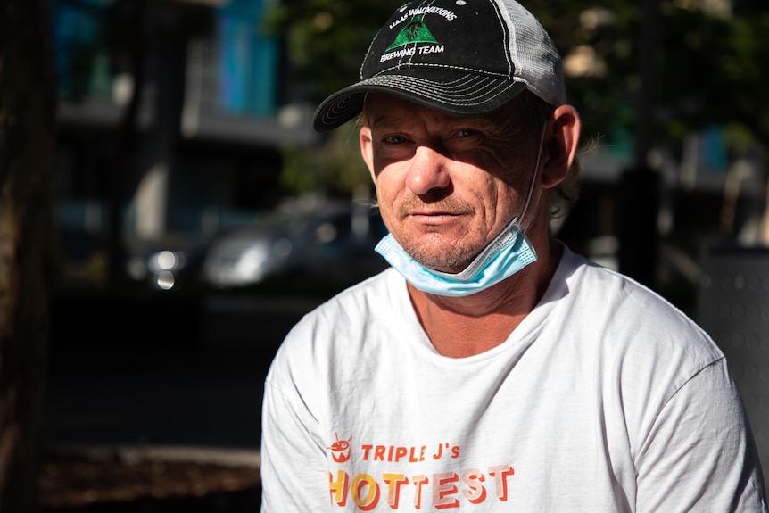 A man wearing a hat and a white t-shirt and a blue surgical mask under his chin sits outside.