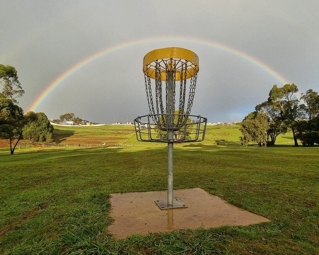 A disc golf hole or metal basket with draping chains and a full rainbow arch behind