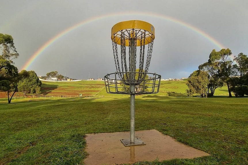 A disc golf hole or metal basket with draping chains and a full rainbow arch behind
