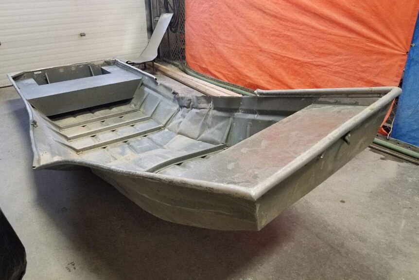 An aluminium boat, bent upwards in the middle, sitting on a concrete floor.