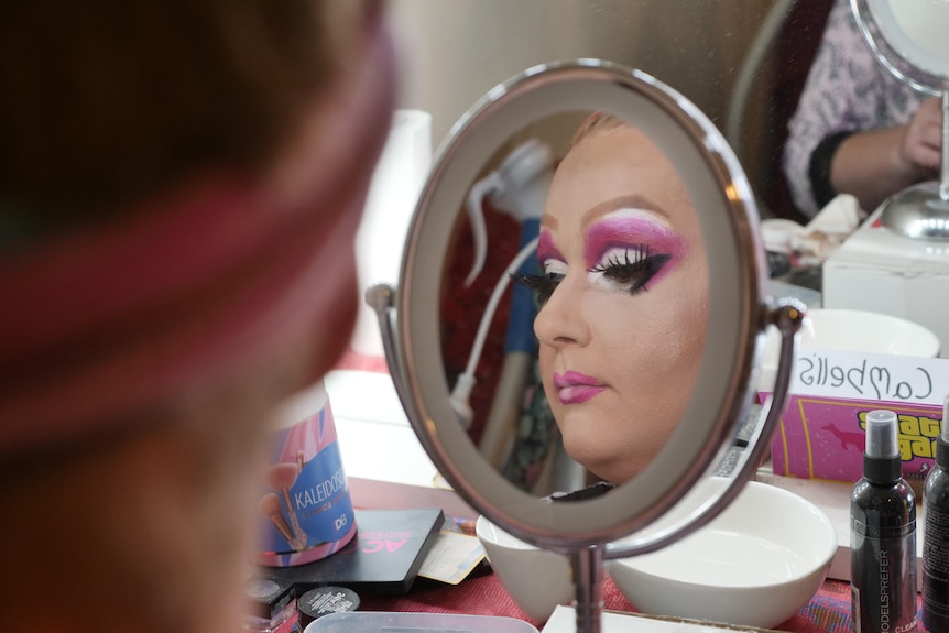 A man wearing bright pink eyeshadow and lipstick looks into a small circle mirror on a make-up desk.