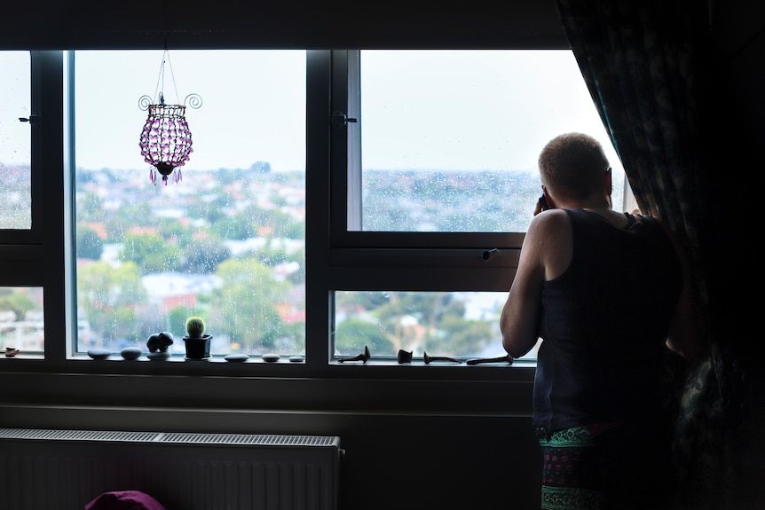 Steph with blonde shirt hair and a blue singlet staring out the window of her room in a high-rise housing commission building.