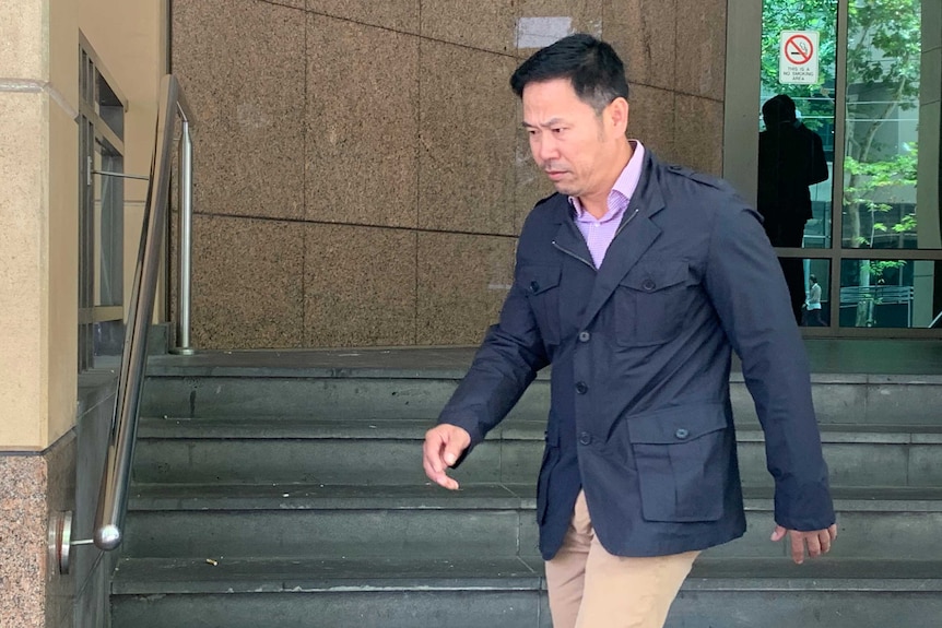 A man with black hair wearing a dark blue jacket and light coloured pants walks down the steps outside court.