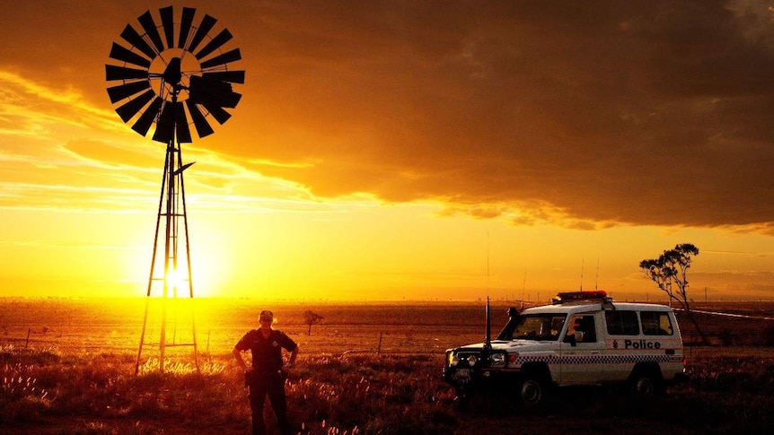 Female police officer stands next to a police vehicle in front of a windmill in a rural setting