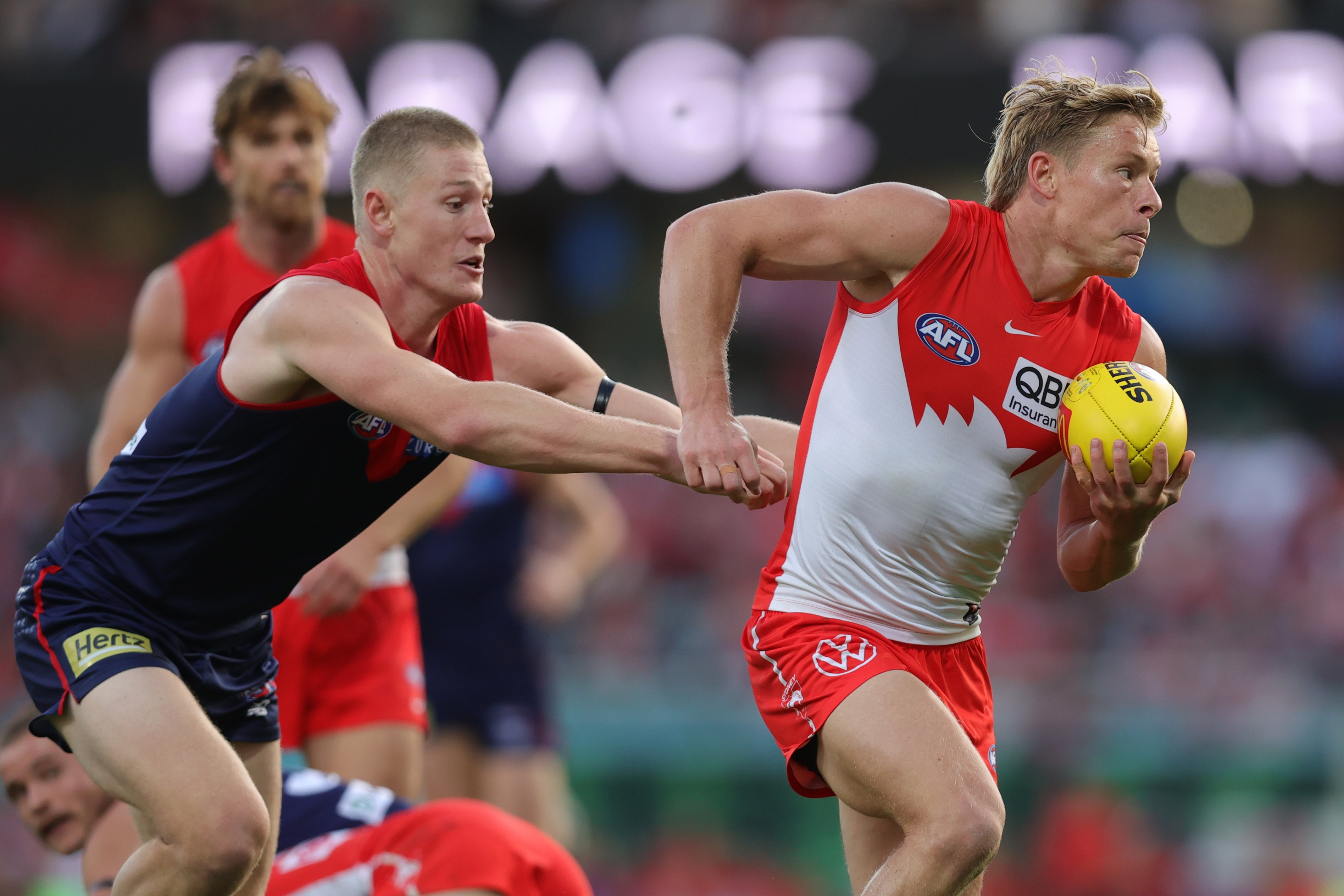 An AFL player in red and white holds a yellow ball, being tackled by a player in blue and red