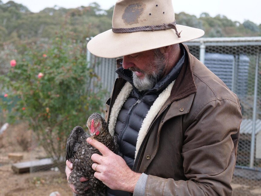 A man wearing a brown jacket and akubra hat holds a chicken.