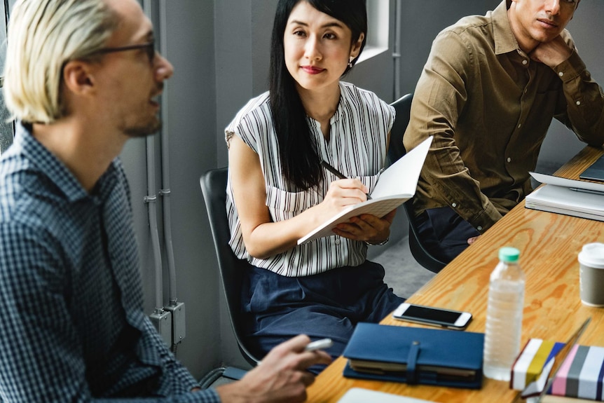 A woman looks at a colleague in a meeting room while writing in a notebook.