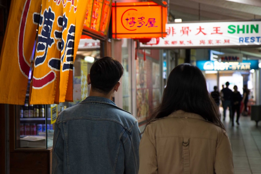 Backs of Mikey and Heidi Tai walking in next to Asian restaurants in the restaurant district of Sunnybank, Brisbane.