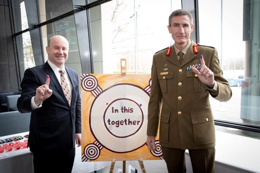 A man in a suit and a man in military uniform holding one finger up next to a sign which says In this together