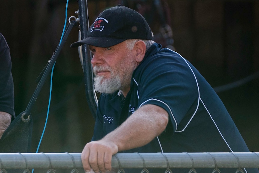 A man with a cap and white beard puts his hand on a fence.