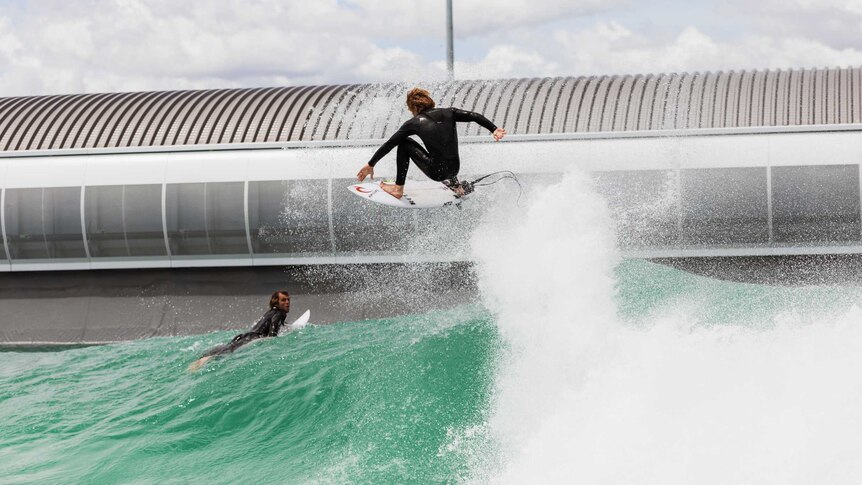 Two surfers, one lying on his surfboard and other wave jumping on top of a blue-green wave with a modern building behind them.