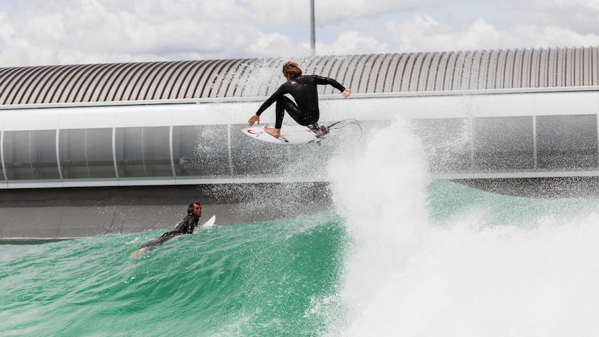 Two surfers, one lying on his surfboard and other wave jumping on top of a blue-green wave with a modern building behind them.