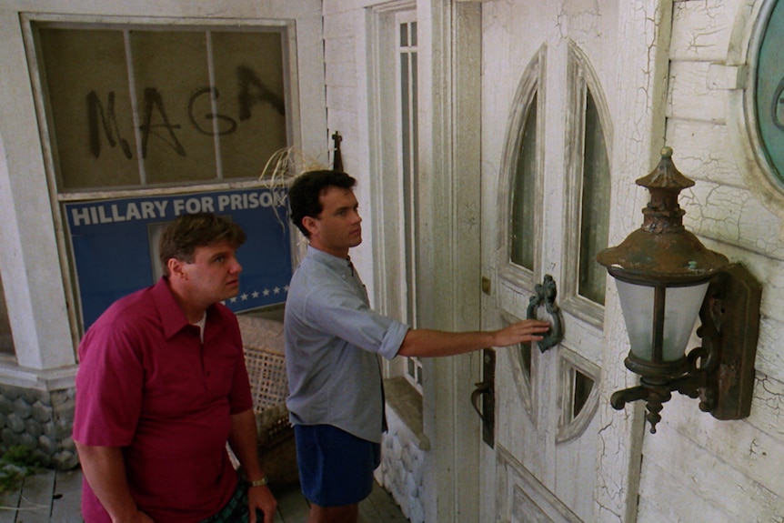 A film still from The 'Burbs, of two men approaching a dilapidated door. In the window behind them MAGA is written in dust.