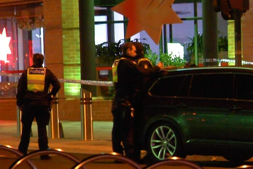 Three police officers stand at the rear of a car at a murder scene at night.