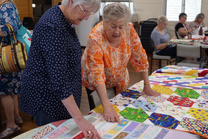 Del Clark in a bright orange patterned shirt looks over some colourful quilts with another Stitch and Chat member.