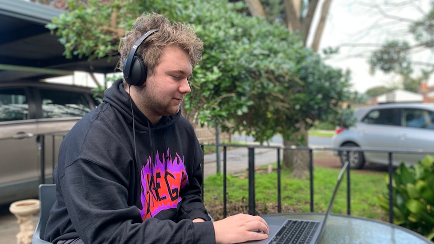 A young boy wears a grey jumper with a purple logo, headphones and works on a laptop, sitting in the open.