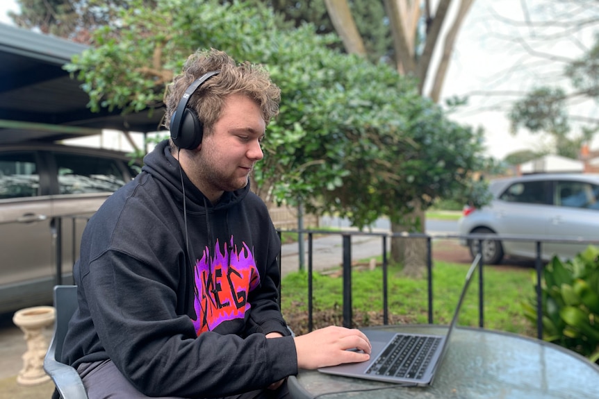 A young boy wears a grey jumper with a purple logo, headphones and works on a laptop, sitting in the open.
