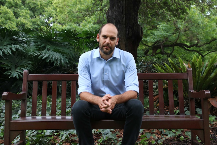 A bearded man sits on a wooden bench with a backdrop of lush vegetation.