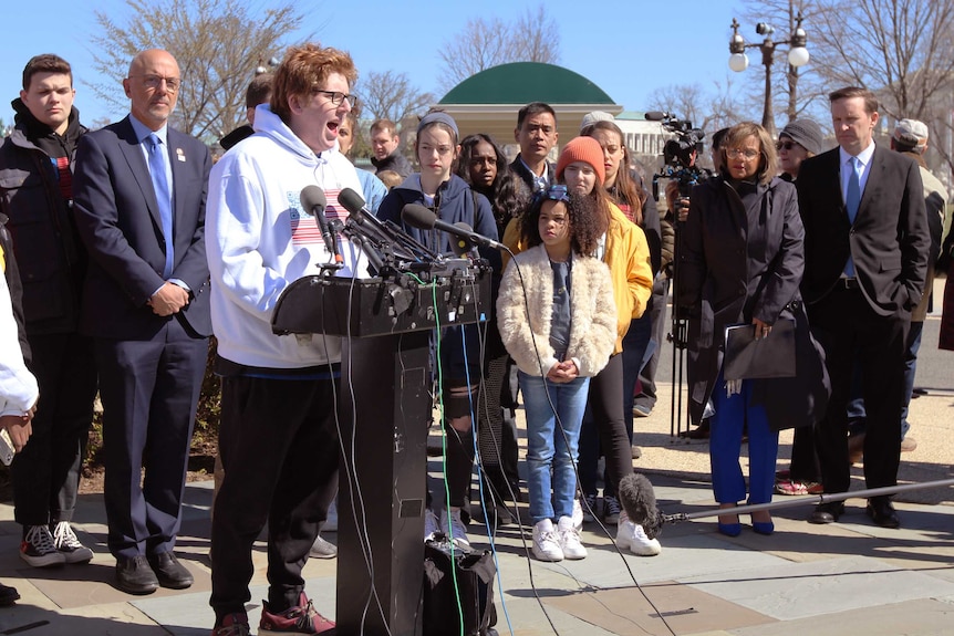 A student speaks into microphones at a lectern as people listen.