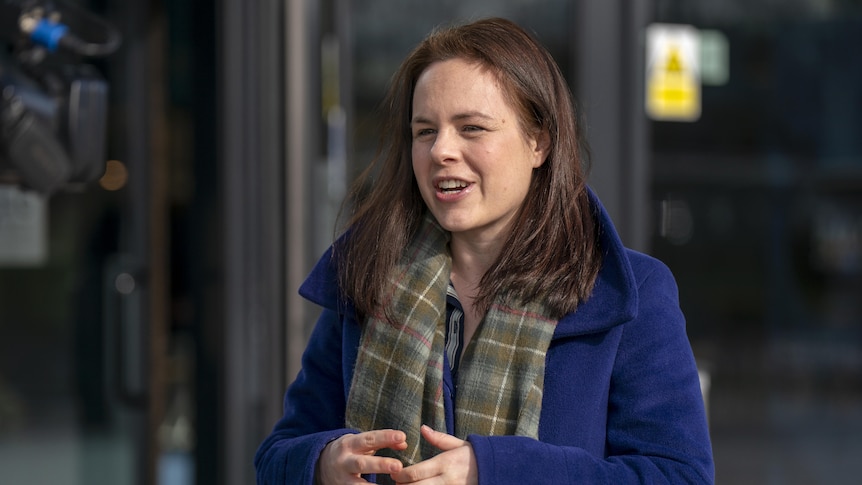 candid image of kate forbes outdoors in blue trench coat and green scarf