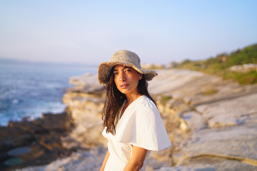Vanessa Conrad stands on a rocky coastal outcrop at sunset, wearing a clothing hemp hat and a white hemp d